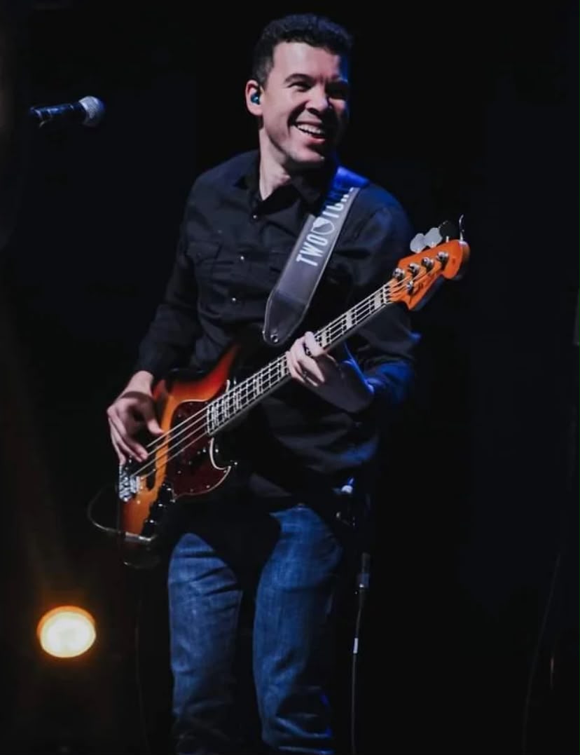Samuel sorrindo tocando baixo no palco, ao lado do microfone